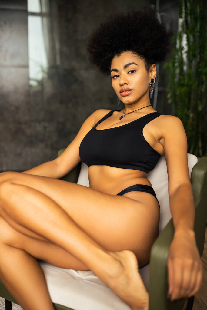 Sexy african american woman in black underwear looking at camera while sitting on armchair at home  - Photo, Image