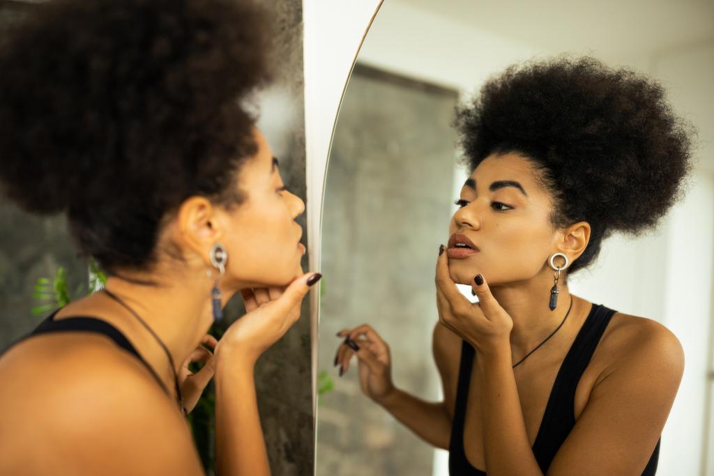 Oldalnézet elmosódott afro-amerikai nő megható arc, miközben nézi a tükröt a fürdőszobában - Fotó, kép
