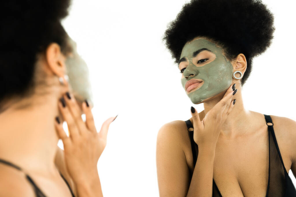 Yüzünde kil maske olan kıvırcık Afrikalı Amerikalı kadın aynaya bakıyor.  - Fotoğraf, Görsel
