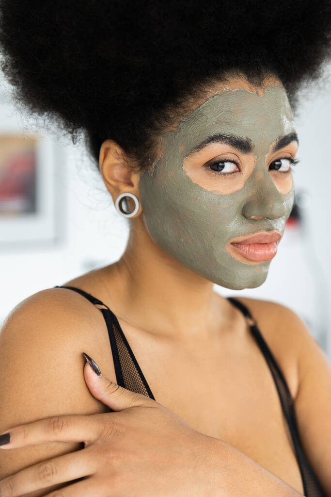 Yüzünde kil maske olan Afro-Amerikan kadın portresi. Evinde kameraya bakıyor.  - Fotoğraf, Görsel