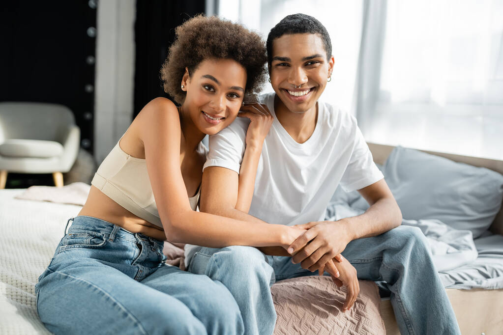 joven y feliz afroamericano pareja en jeans sentado en la cama y mirando a la cámara - Foto, imagen
