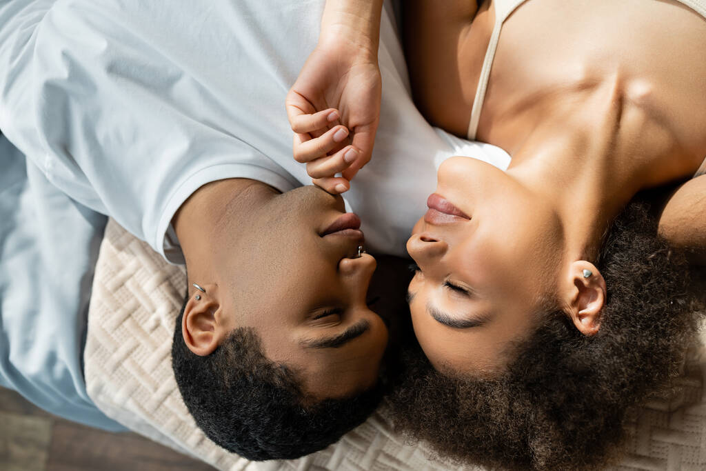 Вид сверху африканской женщины с закрытыми глазами, трогающей подбородок молодого бойфренда на кровати - Фото, изображение