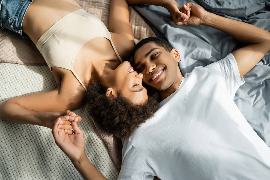 Вид сверху на молодую улыбчивую африканскую пару, лежащую на кровати и держащуюся за руки - Фото, изображение