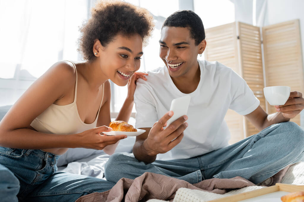 młody Afroamerykanin człowiek z filiżanką kawy pokazując smartfon do uśmiechniętej dziewczyny z rogalikiem w sypialni - Zdjęcie, obraz