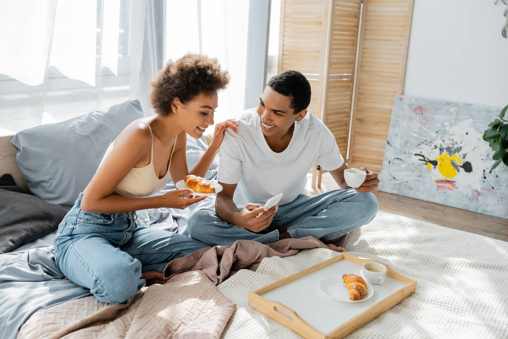 souriant homme afro-américain tenant smartphone et tasse de café près petite amie heureuse avec croissant - Photo, image