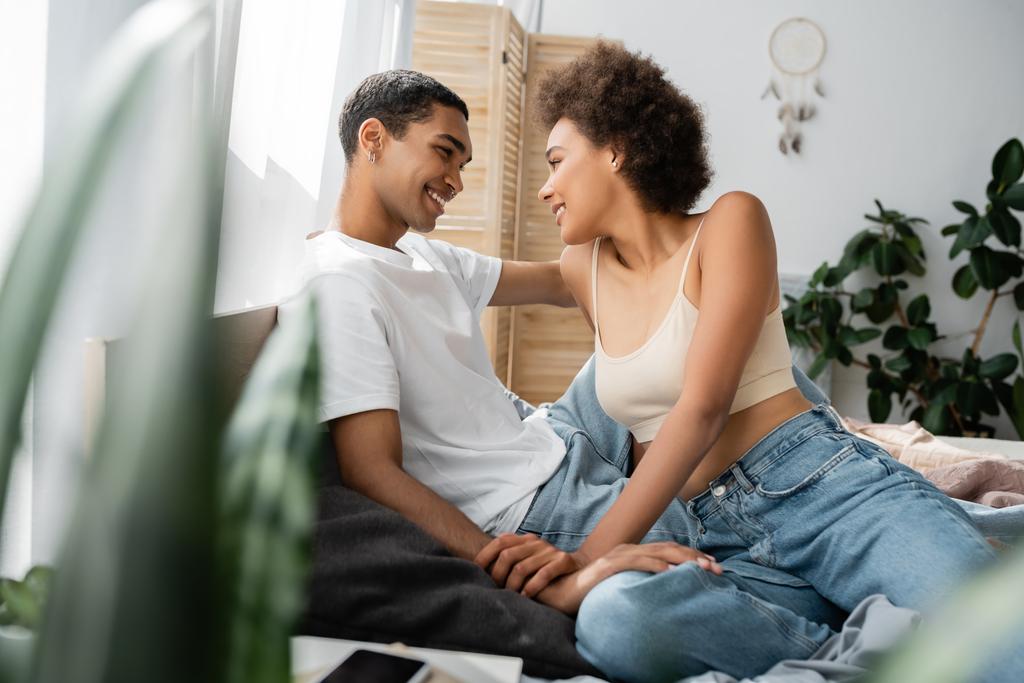sexy couple afro-américain assis sur le lit et souriant à l'autre sur le premier plan flou - Photo, image