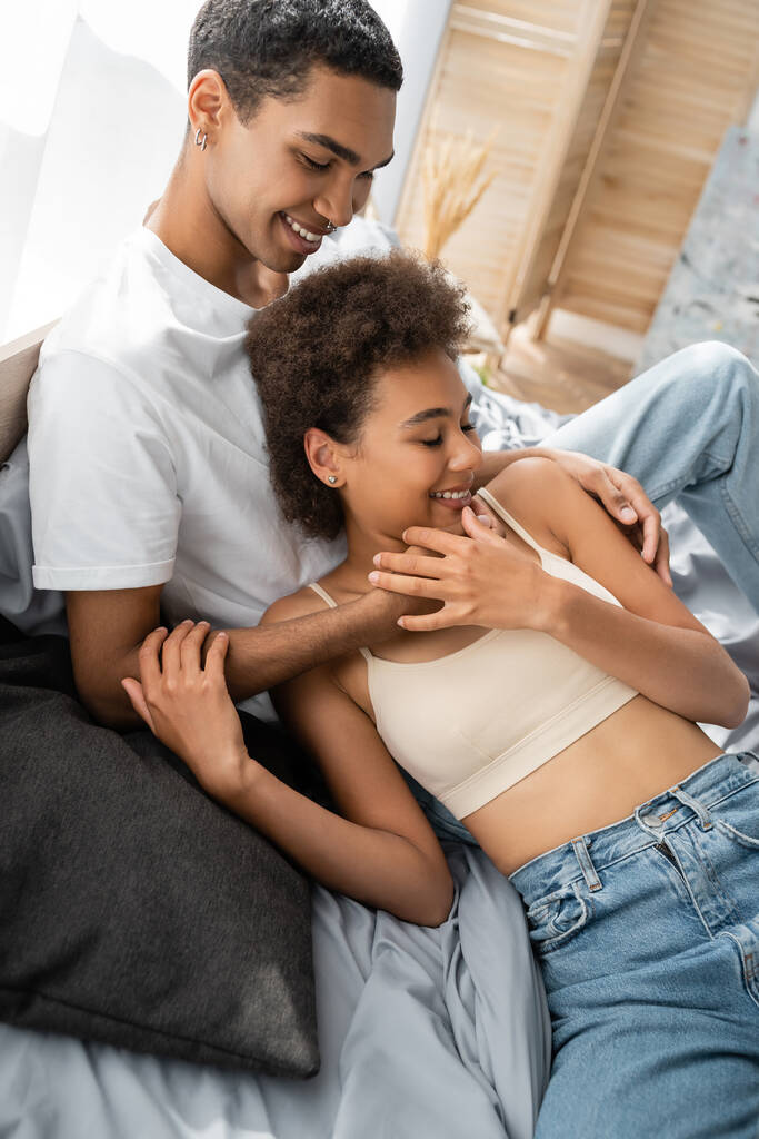 usmívající se africký americký muž objímající sexy přítelkyně v plodině top a džíny na posteli - Fotografie, Obrázek