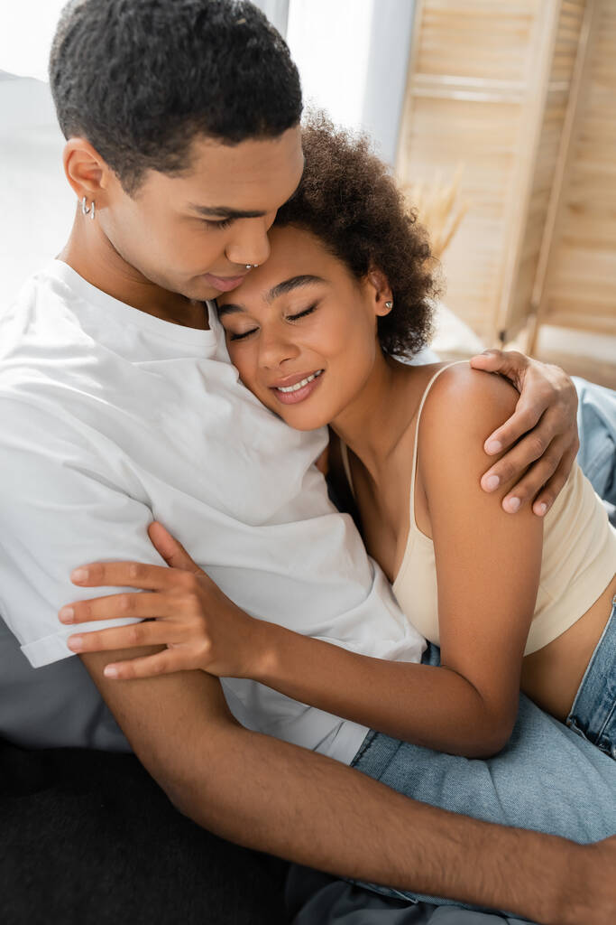 mulher bonita e encaracolada com os olhos fechados abraçando com o namorado afro-americano no quarto - Foto, Imagem