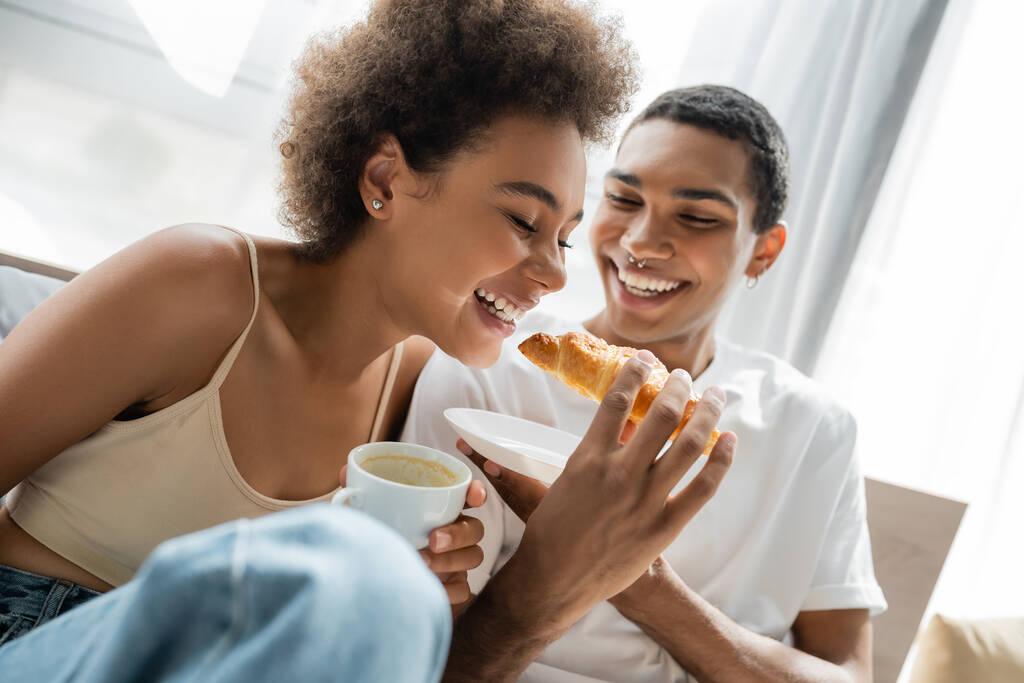 joyful african american man feeding smiling girlfriend with croissant during breakfast in bedroom - Photo, Image