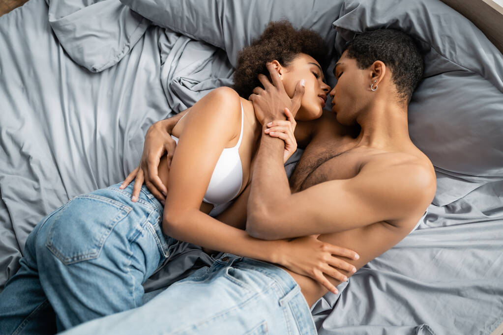 top view of shirless African American mężczyzna przytula gorącą kobietę w dżinsach i stanik na łóżku - Zdjęcie, obraz