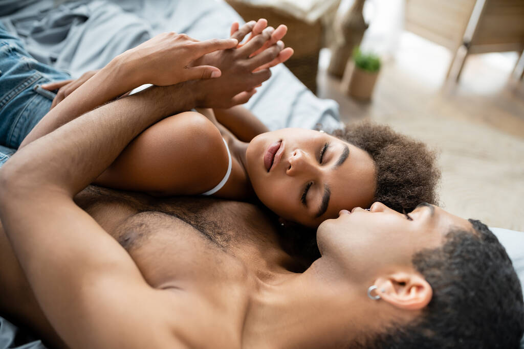 pretty african american woman with closed eyes holding hands with shirtless boyfriend on bed - Photo, Image
