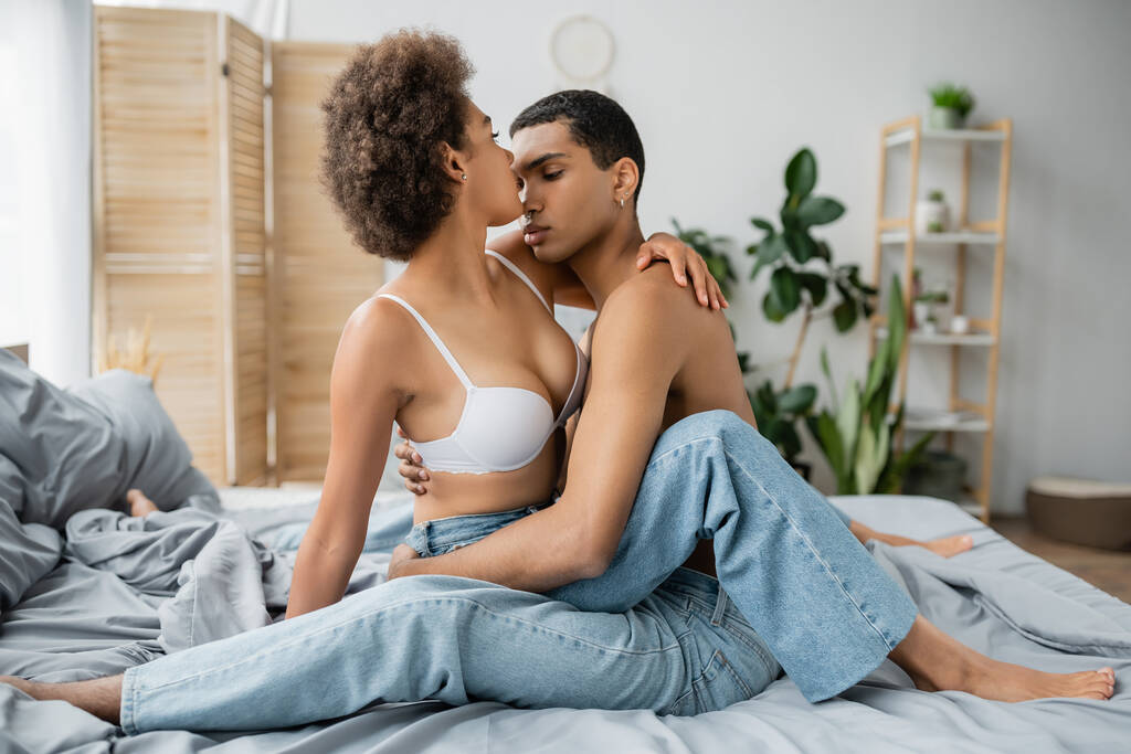 若いですアフリカ系アメリカ人女性とともにセクシー体近いです白人のボーイフレンド座っていますベッドでジーンズ - 写真・画像