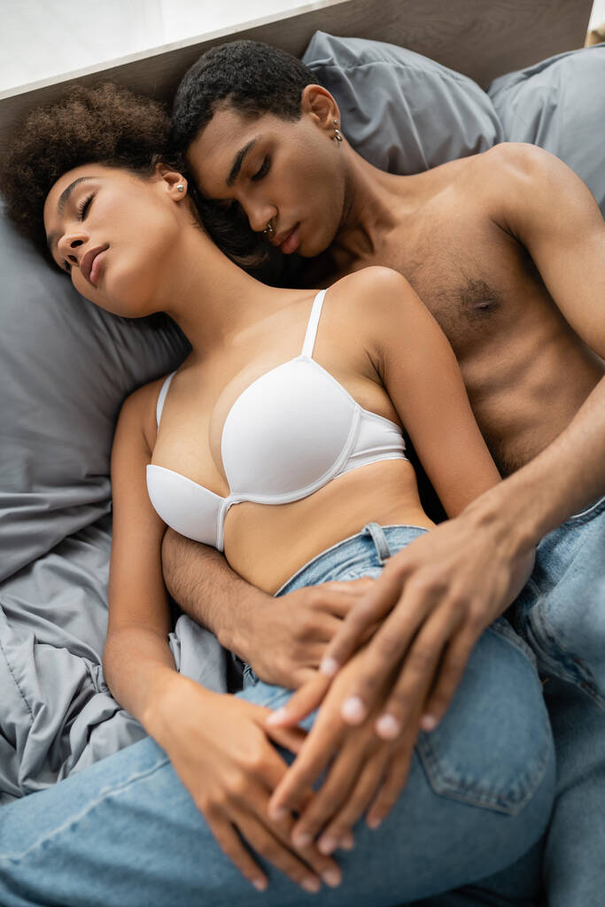 vista de ángulo alto de hombre y mujer afroamericanos sin camisa en sujetador blanco acostado en la cama con los ojos cerrados - Foto, Imagen
