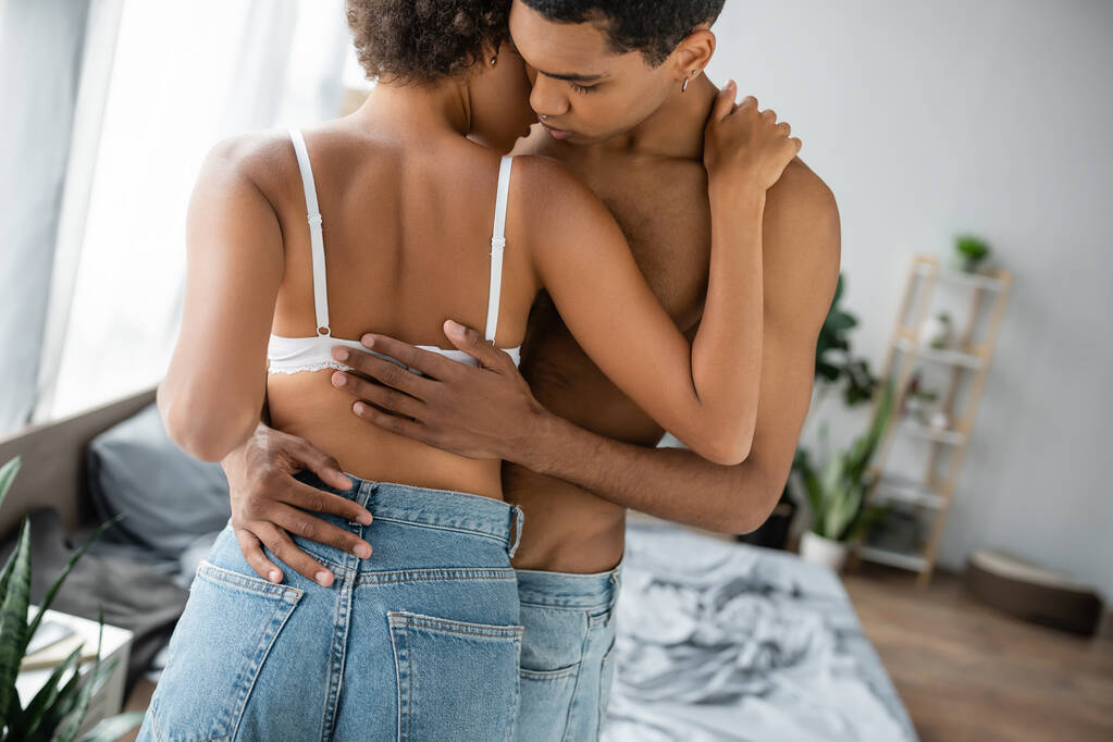 Genç ve üstsüz Afro-Amerikan erkeği. Kotlu ve sütyenli baştan çıkarıcı kız arkadaşı kucaklıyor. - Fotoğraf, Görsel