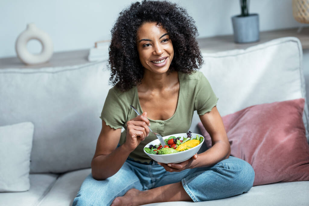 Aufnahme einer schönen Frau, die gesunden Salat isst, während sie zu Hause auf der Couch sitzt. - Foto, Bild