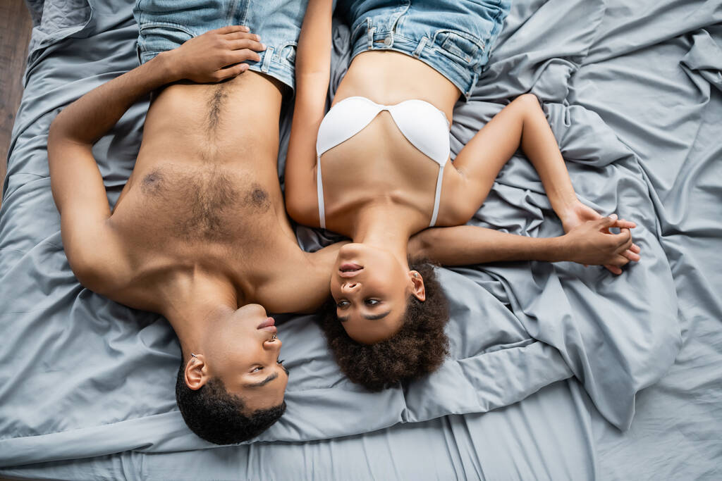 vista dall'alto degli amanti afro-americani sexy che si tengono per mano e si guardano l'un l'altro su biancheria da letto grigia - Foto, immagini