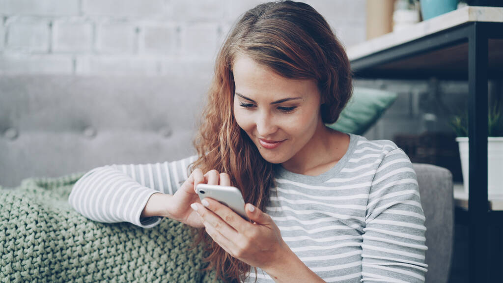 Onnellinen nuori nainen pitää älypuhelinta kosketusnäytöllä ja hymyilee tarkistamalla sosiaalisen median tilin tai lukemalla tekstiviestejä. Moderni teknologia ja millennials käsite
. - Valokuva, kuva