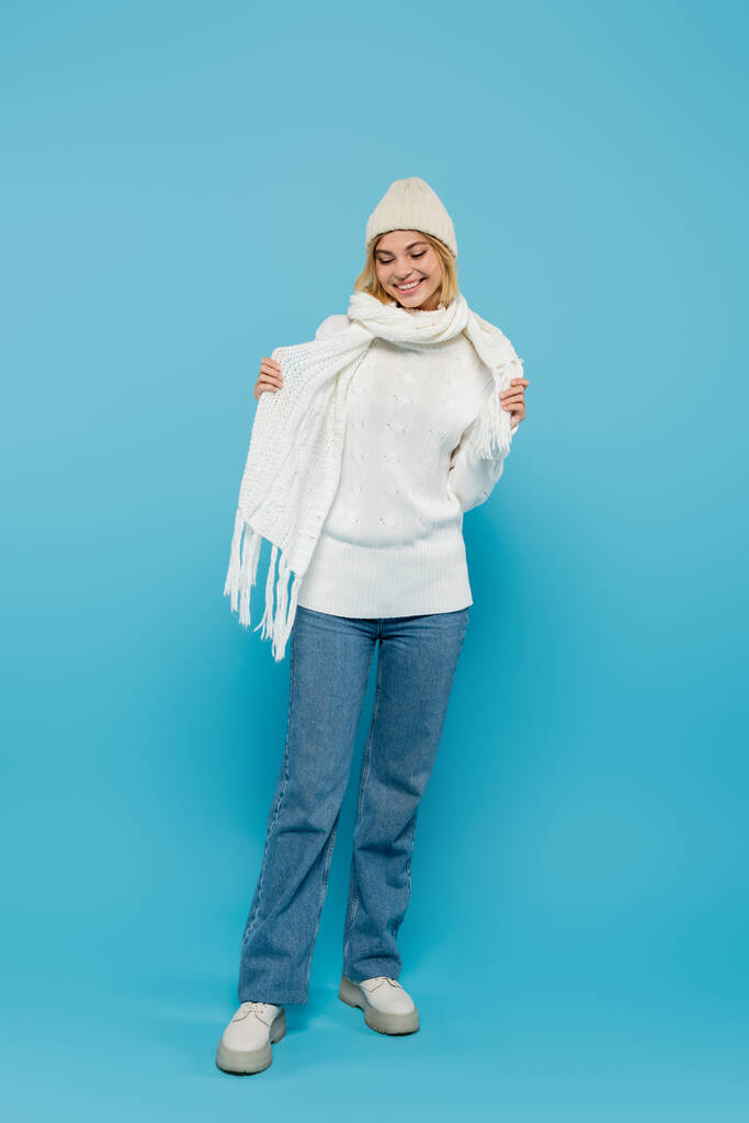 longitud completa de mujer rubia alegre en suéter blanco y sombrero ajustando bufanda de invierno en azul - Foto, Imagen