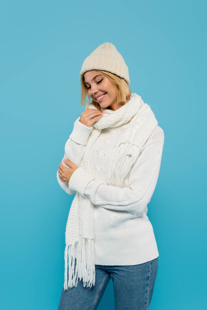 szczęśliwa blondynka kobieta w białym swetrze i zimowym kapeluszu uśmiechnięta odizolowana na niebiesko  - Zdjęcie, obraz