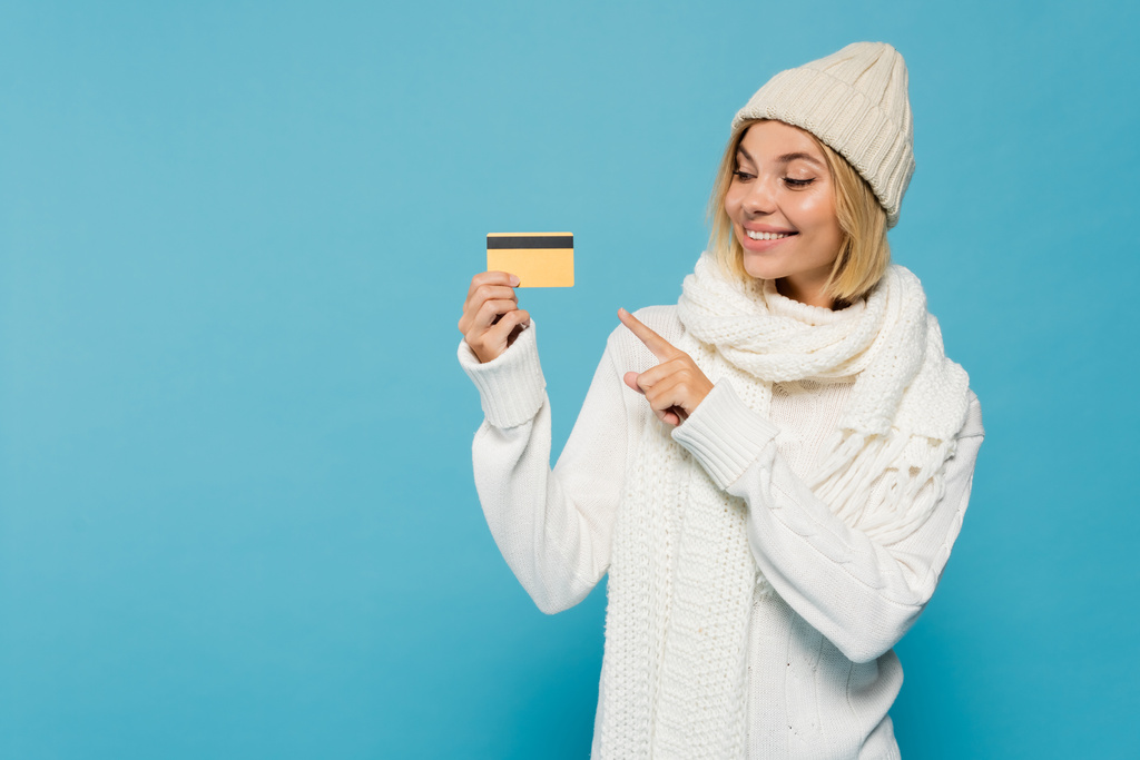 happy blonde woman in white sweater and winter hat pointing with finger at credit card isolated on blue - Photo, Image