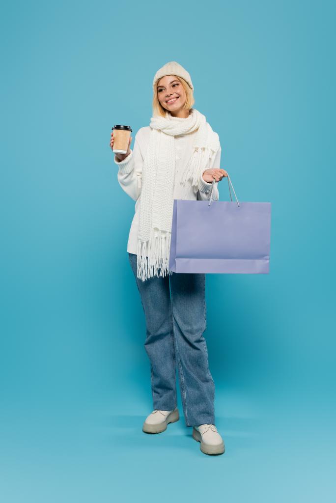 full length of happy blonde woman in white sweater and knitted hat holding paper cup and shopping bag on blue - Photo, Image