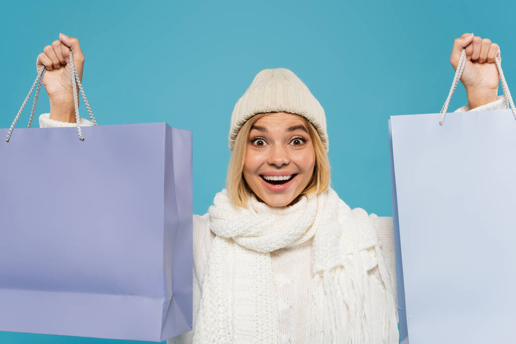 白いセーターとニット帽を着た興奮した若い女性青で隔離されたショッピングバッグを保持 - 写真・画像