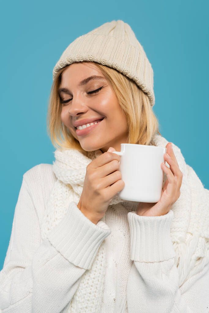 elégedett fiatal nő fehér pulóver és kalap gazdaság csésze kávé elszigetelt kék  - Fotó, kép
