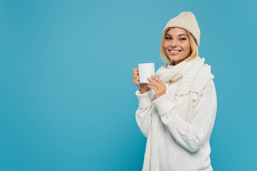радостная молодая женщина в белом свитере и шляпе держа чашку кофе изолированы на синий  - Фото, изображение