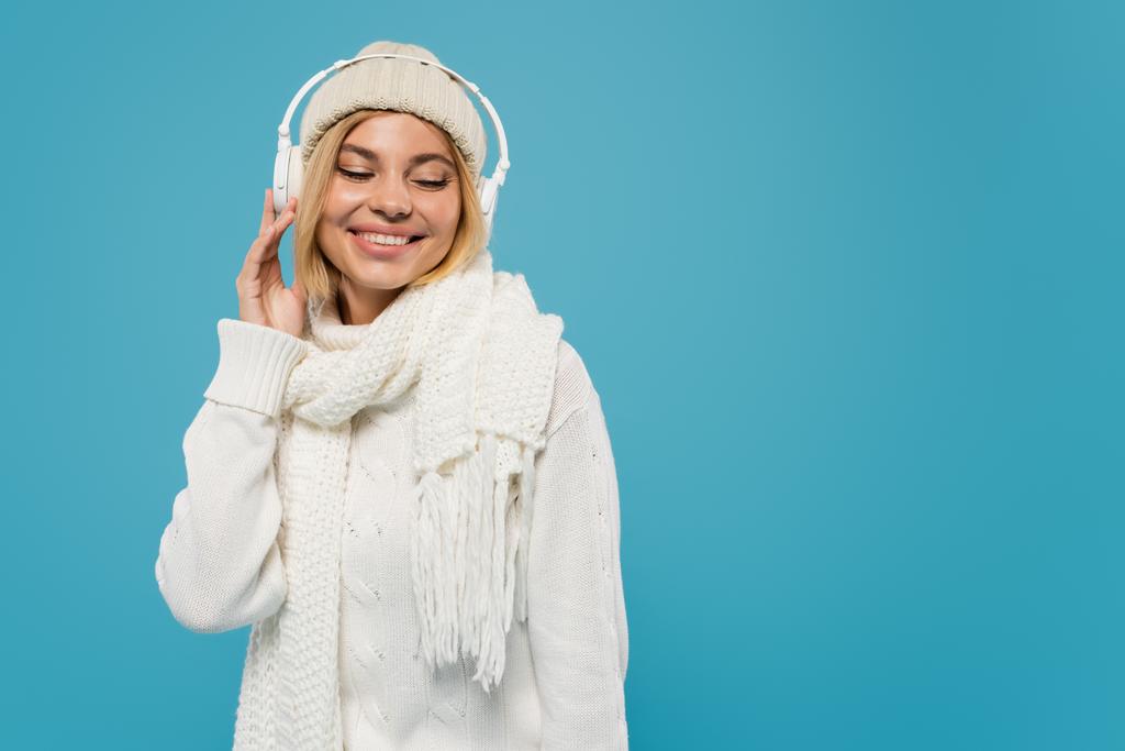χαμογελαστή γυναίκα σε λευκό πουλόβερ και καπέλο ακούγοντας μουσική σε ασύρματα ακουστικά που απομονώνονται σε μπλε  - Φωτογραφία, εικόνα