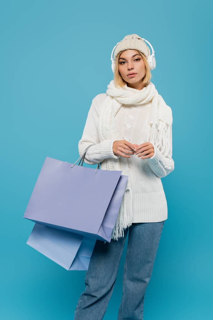 mujer joven en traje de invierno y auriculares inalámbricos que sostienen bolsas de compras aisladas en azul  - Foto, imagen