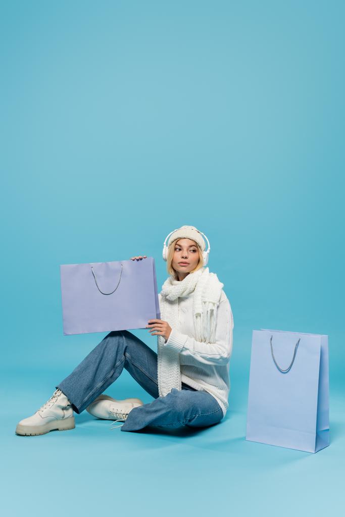 longitud completa de la mujer rubia en traje de invierno y auriculares inalámbricos sentado y sosteniendo la bolsa de compras en azul  - Foto, Imagen