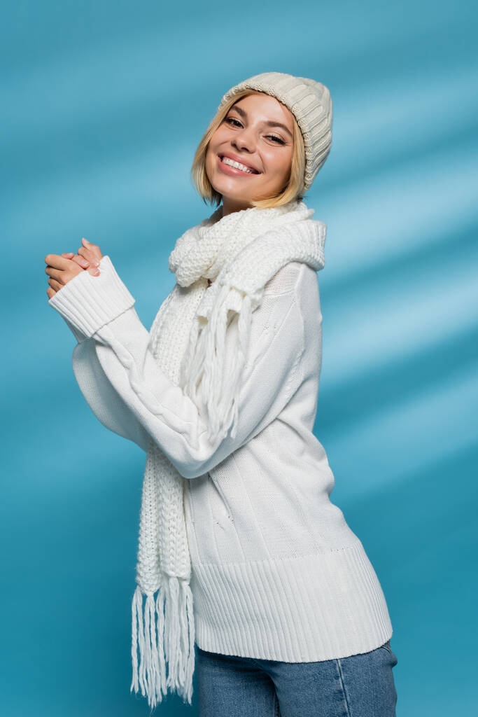 szczęśliwa młoda kobieta w dzianinie kapelusz i zimowy sweter uśmiecha się na niebiesko  - Zdjęcie, obraz
