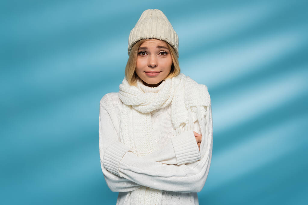 pretty young woman in knitted hat and winter sweater feeling cold while standing with crossed arms on blue  - Photo, Image