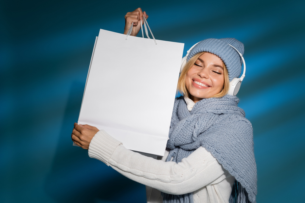 задоволена молода жінка в зимовому шарфі і капелюсі слухає музику в бездротових навушниках і тримає сумку на синьому
  - Фото, зображення