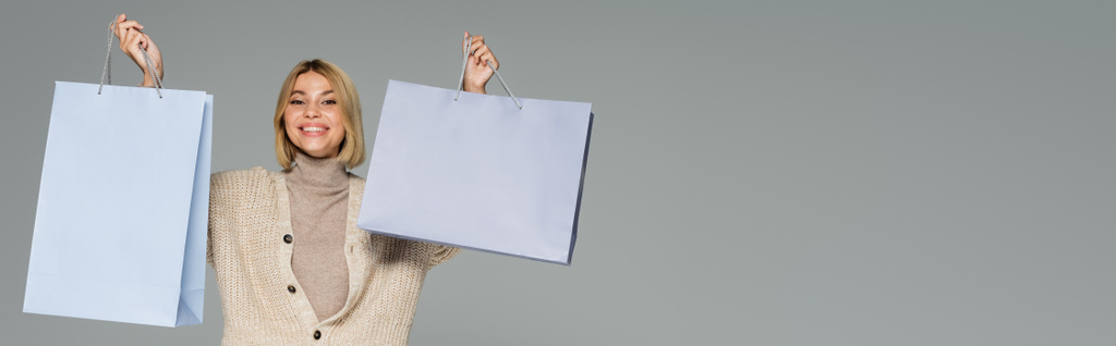 boldog szőke nő garbó és kardigán gazdaság bevásárló táskák elszigetelt szürke, banner - Fotó, kép