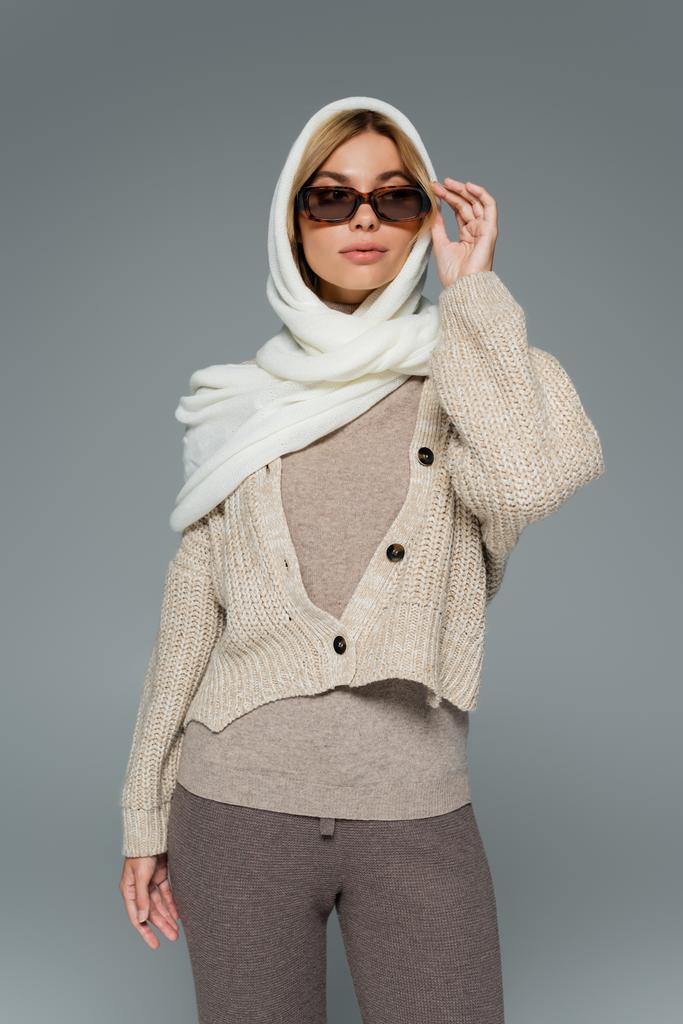 jonge vrouw in gebreide kleding en hoofddoek aanpassen zonnebril geïsoleerd op grijs - Foto, afbeelding