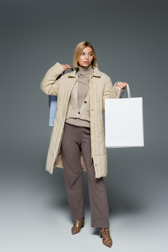Mujer de moda en chaqueta de invierno y cuello alto sosteniendo bolsas de compras sobre fondo gris  - Foto, Imagen
