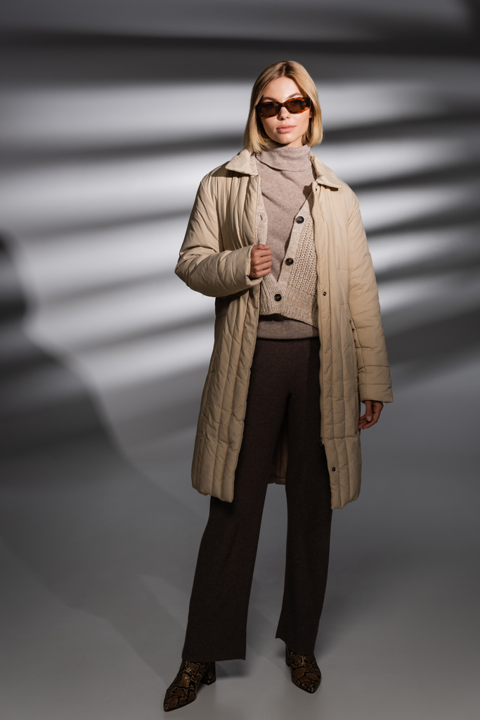 Piena lunghezza di donna alla moda in giacca invernale e occhiali da sole in piedi su sfondo grigio astratto  - Foto, immagini