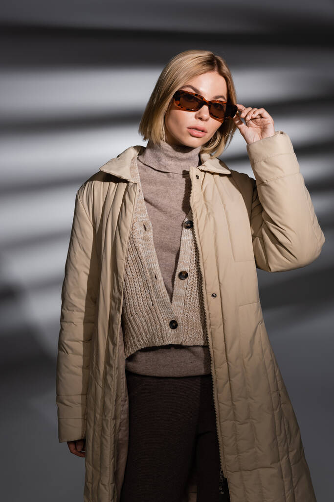 Modna młoda kobieta w okularach przeciwsłonecznych i zimowej kurtce pozująca na abstrakcyjnym szarym tle  - Zdjęcie, obraz