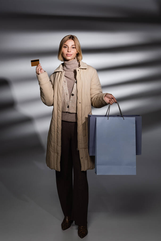 Giovane donna in giacca invernale in possesso di carta di credito e shopping bag su sfondo grigio astratto  - Foto, immagini
