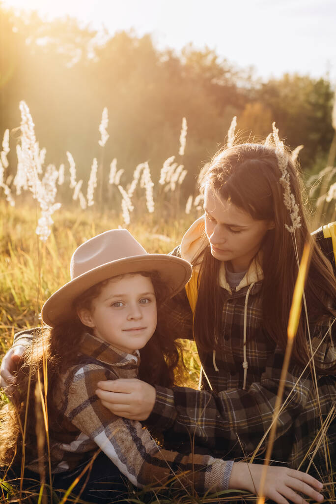 Retrato de sonriente adorable hijita en sombrero con hermosa madre joven sentada en la hierba sobre el fondo hermoso bosque de otoño durante la increíble puesta de sol. Concepto relaciones familiares y viajes - Foto, Imagen