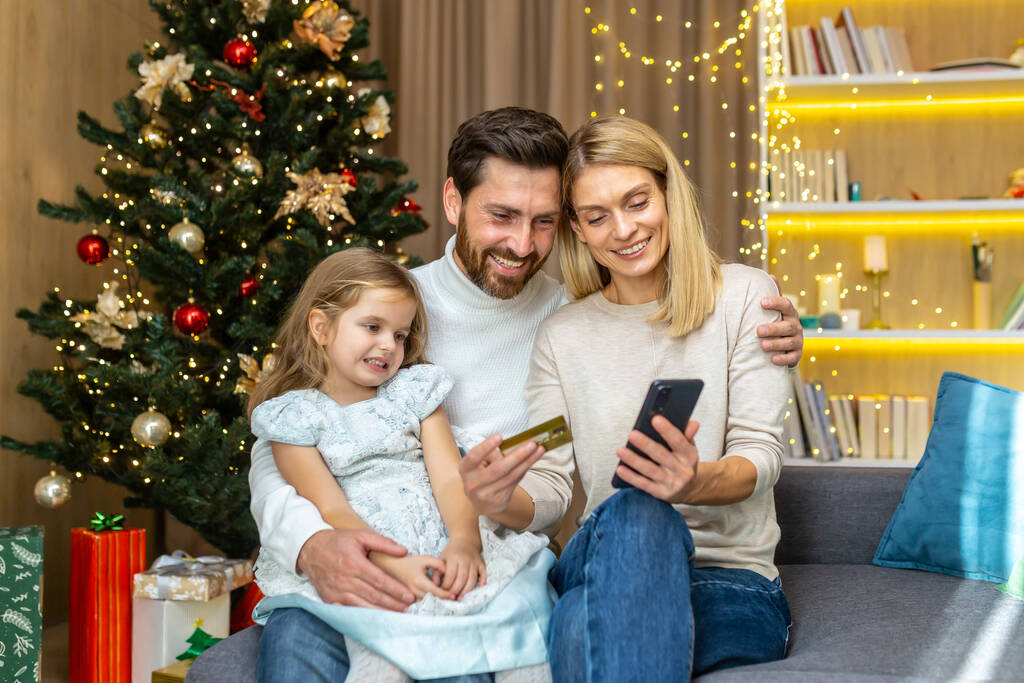 Famiglia felice per lo shopping di Natale online scegliendo regali nel negozio online, uomo e donna con bambino seduti a casa sul divano vicino all'albero di Natale con carta di credito bancaria. - Foto, immagini