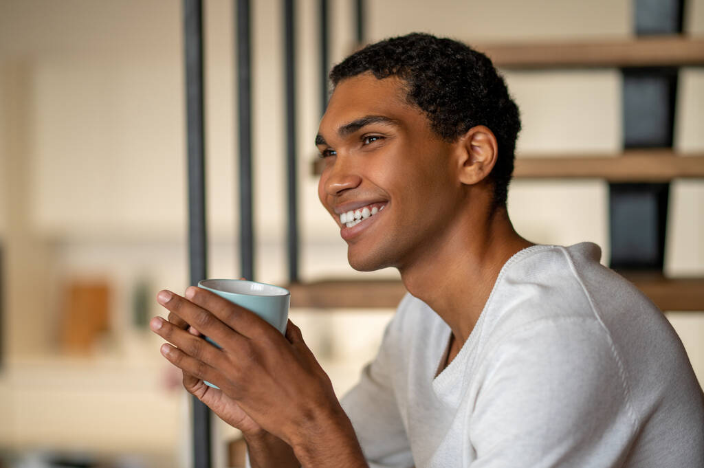 Café da manhã. Jovem homem sorridente em casa tomando café e olhando satisfeito - Foto, Imagem