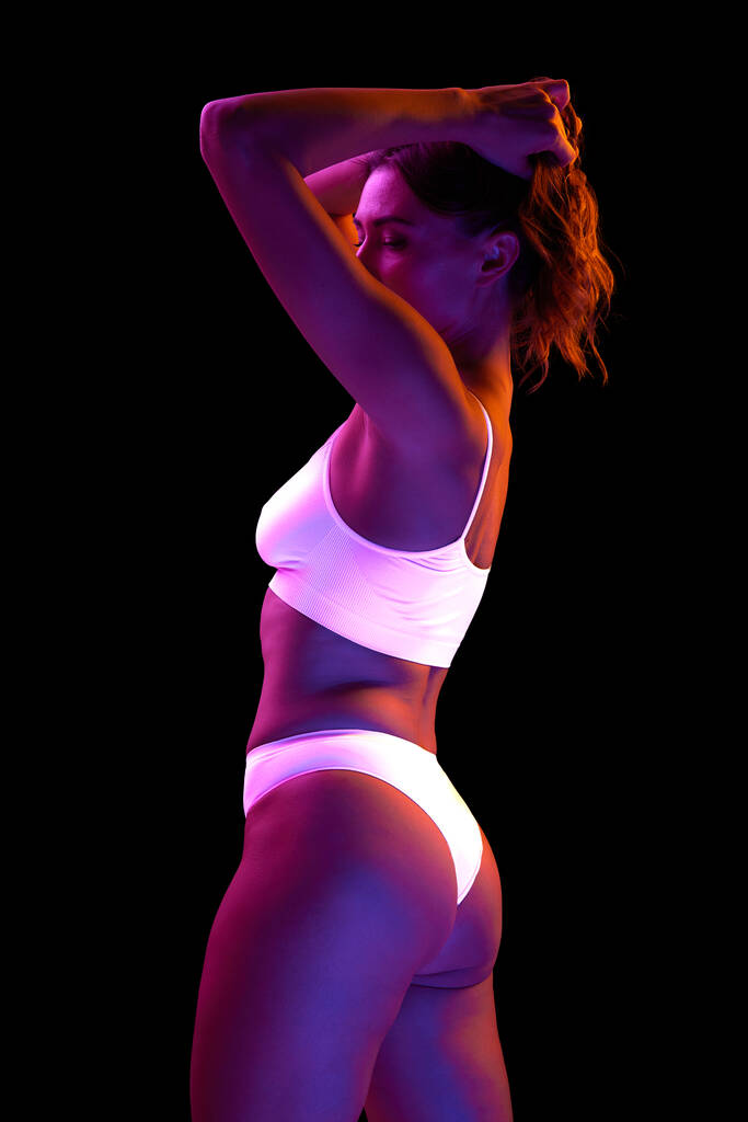 Porträt einer schönen Frau mit schlankem Körper, die in weißer Unterwäsche isoliert vor schwarzem Hintergrund in Neonlicht posiert. Konzept der Schönheit, Körper- und Hautpflege, Gesundheit, plastische Chirurgie, Kosmetik, Sport - Foto, Bild