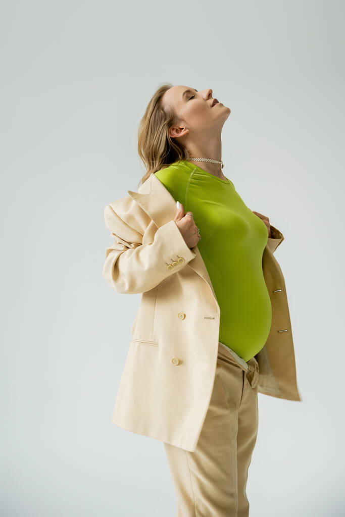 Κομψό έγκυος γυναίκα αγγίζοντας μπεζ σακάκι, ενώ στέκεται απομονωμένο σε γκρι  - Φωτογραφία, εικόνα