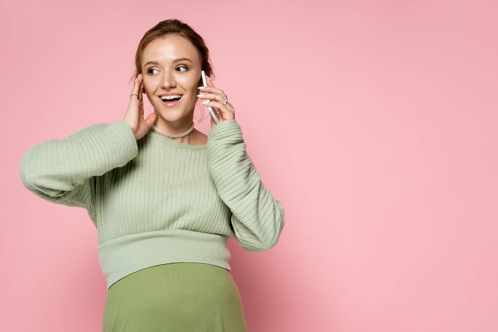 Positiv schwangere Frau in grünem Outfit spricht auf Smartphone auf rosa Hintergrund  - Foto, Bild