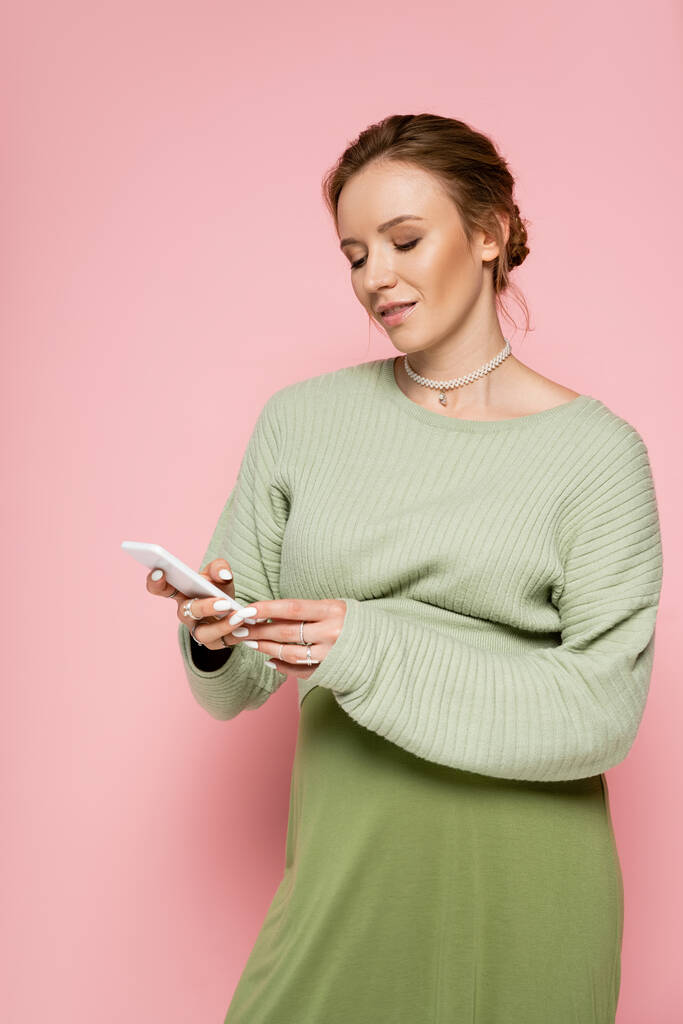 Stylish pregnant woman in green outfit using mobile phone on pink background  - Photo, Image