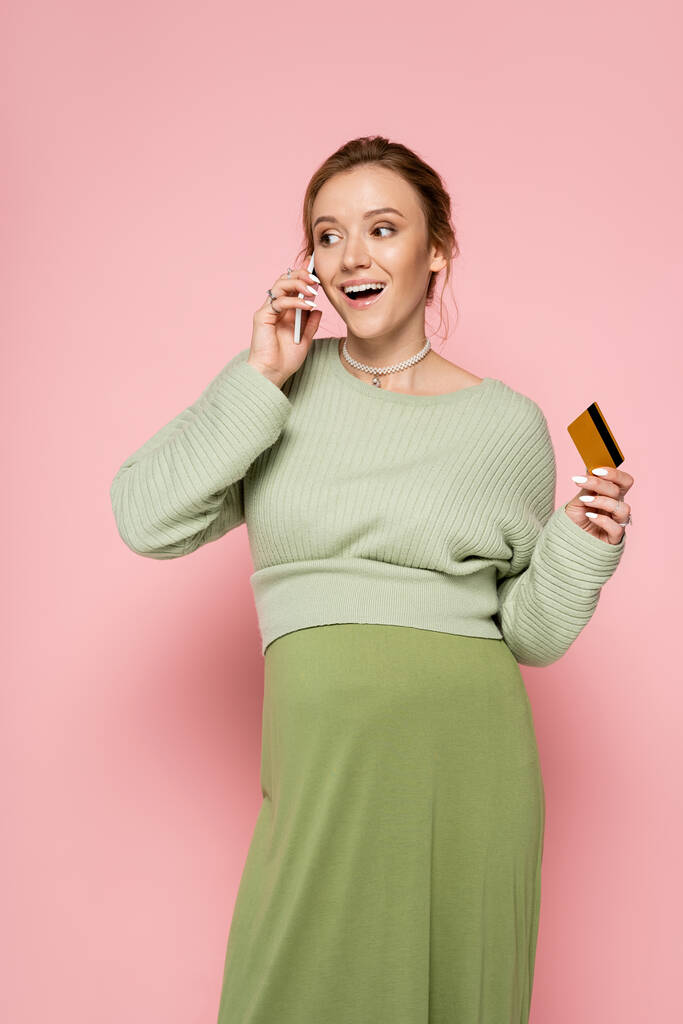 Mujer embarazada emocionada en traje verde con tarjeta de crédito y hablando en el teléfono inteligente sobre fondo rosa  - Foto, Imagen