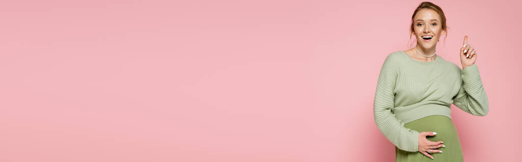 Positive Schwangere im Pullover zeigt mit Finger auf rosa Banner  - Foto, Bild