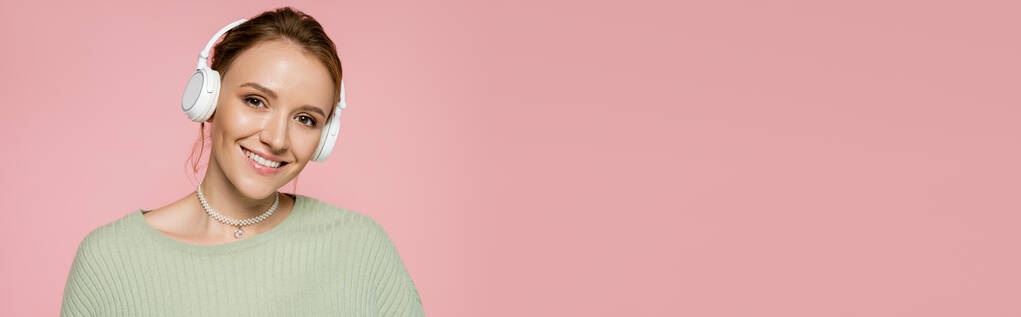 Positive Frau mit Kopfhörer und grünem Pullover blickt vereinzelt in die Kamera auf rosa Banner  - Foto, Bild